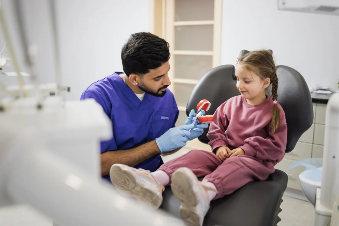 A dentist in scrubs demonstrates dental care using a model to a young girl seated in a dental chair.